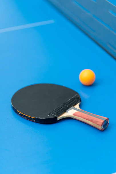 Μια ρακέτα του τένις και μια πορτοκαλί μπάλα του τένις βρίσκονται σε ένα μπλε τραπέζι του τένις δίπλα σε ένα δίχτυ σε ένα πάρκο της πόλης γκρο πλαν του πινγκ πονγκ παιχνίδι - Φωτογραφία, εικόνα