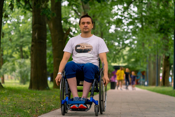 Ένταξη Ένας άνδρας με αναπηρίες οδηγεί μια αναπηρική καρέκλα κατά μήκος του μονοπατιού του πάρκου της πόλης με φόντο τα δέντρα - Φωτογραφία, εικόνα
