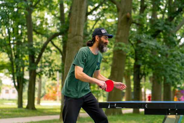 Ein älterer Mann mit Mütze und Schläger spielt in einem Stadtpark vor der Kulisse von Bäumen Tischtennis - Foto, Bild