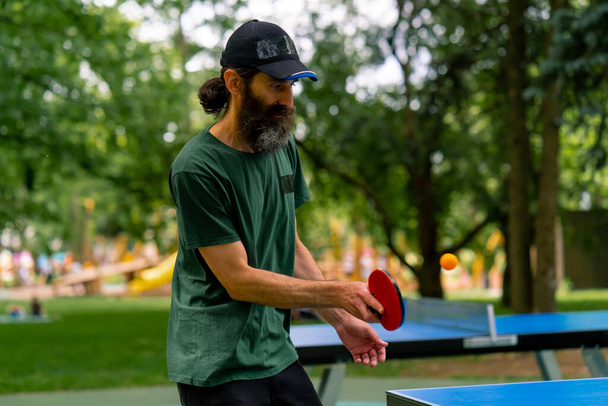 Un hombre mayor usando una gorra y sosteniendo una raqueta juega ping pong en un parque de la ciudad contra el telón de fondo de los árboles - Foto, Imagen