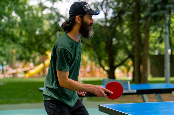 Un hombre mayor usando una gorra y sosteniendo una raqueta juega ping pong en un parque de la ciudad contra el telón de fondo de los árboles - Foto, imagen