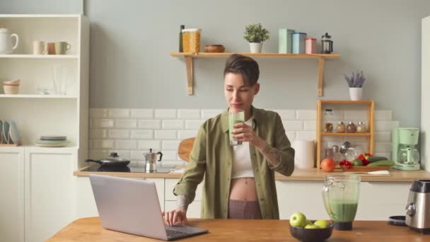 Medium shot of contemporary pregnant woman with short haircut and tattoos using laptop and drinking vitamin green smoothie at kitchen - Filmmaterial, Video