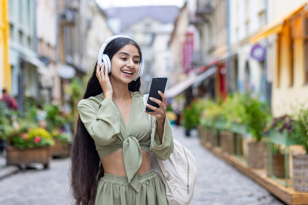 Young beautiful id woman walks in the city, tourist holds phone in hands, uses headphones to listen to music and online radio podcasts, dances and sings along happily. - Photo, image