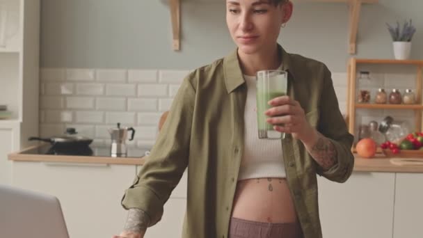 Tilt shot of modern pregnant woman with short dark hair and tattoos on her arms and belly working on laptop from home drinking vitamin green smoothie - Filmati, video