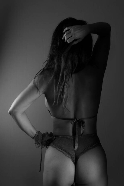 Black and white portrait of sensual model wearing lingerie, standing, making sexy poses. Studio photo. Against dark background. - Photo, Image