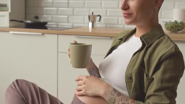 Modern young pregnant woman with short dark hair and tattoos on her arms and belly drinking tea while watching videos on laptop at kitchen table in her apartment - Video
