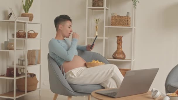 Young Caucasian pregnant woman with short haircut eating junk food while using smartphone sitting in armchair at home - Imágenes, Vídeo