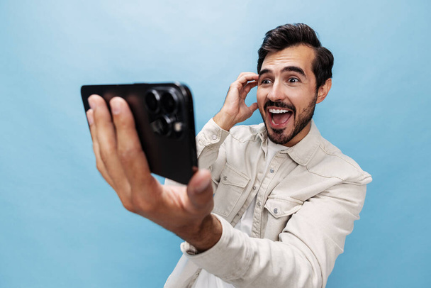 Πορτρέτο ενός κομψού άνδρα μελαχρινή έκπληξη και ανοιχτό στόμα κοιτάζει το blogger τηλέφωνο με γενειάδα, σε ένα μπλε φόντο σε ένα λευκό T-shirt και τζιν, αντίγραφο χώρου. Υψηλής ποιότητας φωτογραφία - Φωτογραφία, εικόνα