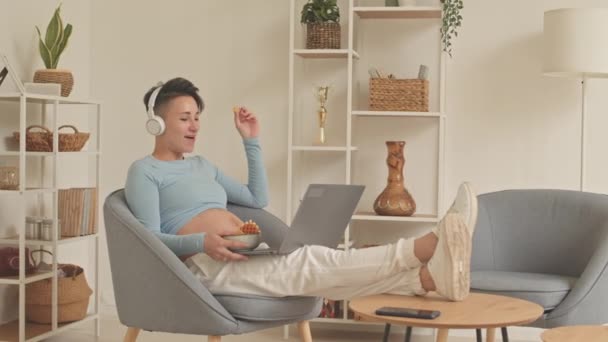 Joyful young pregnant woman in headphones watching comedy film on laptop and eating unhealthy sweets - Metraje, vídeo