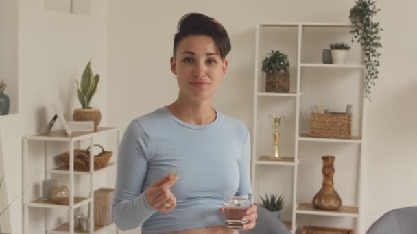 Medium portrait of young short haired pregnant woman smiling at camera holding prenatal vitamin pill and glass of water in hands - Filmagem, Vídeo