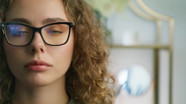 Portrait of attractive female student with curly brunette hair opening eyes and looking to camera in modern apartment. Close up face of beautiful woman in stylish glasses with black modern frame 4K - Séquence, vidéo