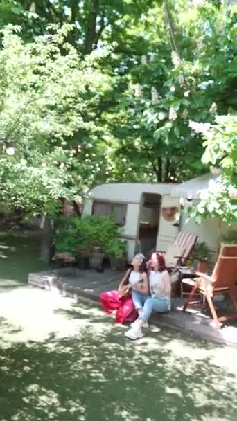 camera zooms in Two beautiful hippie girls are singing a song sitting on the terrace near the trailer with a guitar. High quality 4k footage - Materiaali, video