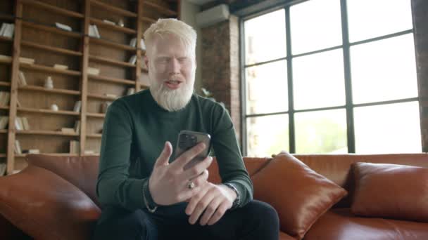 Discolored white haired man with albinisms disease causing neurological disorder with involuntary head shaking trembling. Portrait of Albino guy taking by video call using front camera on smartphone - Кадры, видео