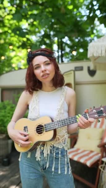 The beautiful hippie girl is playing the guitar and singing near the trailer. High quality 4k footage - Záběry, video