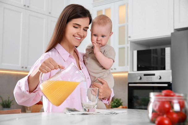 Mother holding her cute little baby while pouring juice into glass in kitchen, space for text - Foto, afbeelding
