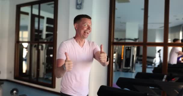 Cheerful man shows thumbs up running on treadmill in modern gym. Concept of sports club endorsement and body shape maintenance slow motion. Healthy lifestyle - Filmmaterial, Video