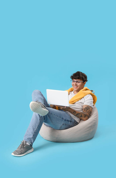 Jeune homme utilisant un ordinateur portable dans un sac de fèves sur fond bleu - Photo, image