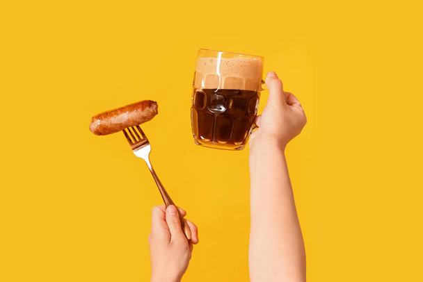 Женские руки с кружкой холодного пива и колбасой на желтом фоне - Фото, изображение