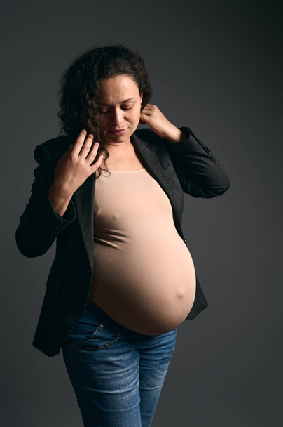 Authentic gorgeous adult pregnant woman dressed in a beige bodysuit, stylish gray blazer and blue jeans, posing against a fashion gray studio background. Beautiful pregnancy. Maternity lifestyle - 写真・画像