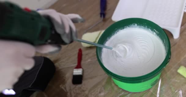 Builder stirs white paint in bucket with industrial mixer on floor in room slow motion. Worker prepares color material for house maintenance - Felvétel, videó