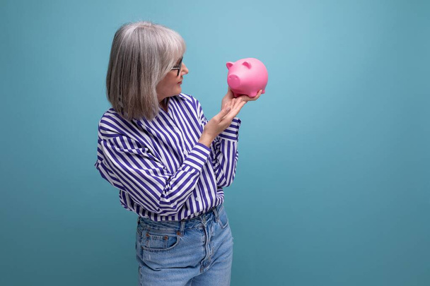 пенсионные накопления. Женщина средних лет с седыми волосами держит копилку с деньгами на ярком фоне студии. - Фото, изображение