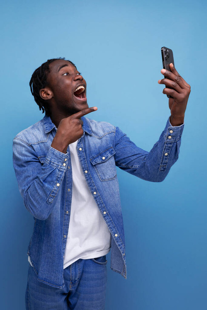 スマートフォンのビデオリンクで話すデニムジャケットでドレッドロックを持つ陽気な若いアフリカ人男性の肖像画. - 写真・画像