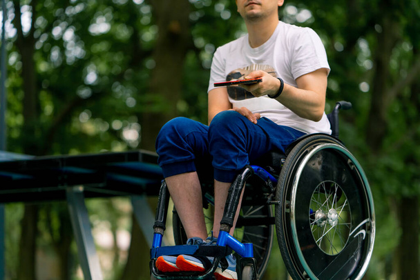 Mavi pin pon masasının yanındaki tekerlekli sandalyedeki özel engelli adam şehir parkındaki tenis raketinde turuncu bir topa vuruyor. - Fotoğraf, Görsel