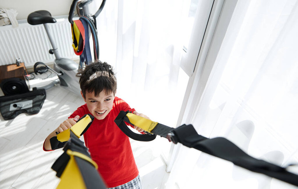 View from above of an adorable Caucasian child, preteen boy exercising with suspension straps indoors. Children. Building Endurance During Bodyweight Training. Sport. Active healthy lifestyle concept - Photo, Image
