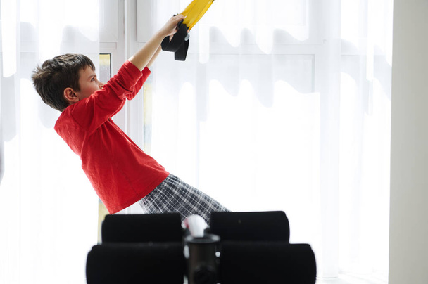Side view of adorable Caucasian child, preteen boy exercising with suspension straps at home. Children. Building Endurance During Bodyweight Training. Sport. Fitness. Active healthy lifestyle concept - Foto, imagen