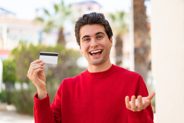 Jonge knappe man met een creditcard in de buitenlucht met geschokte gezichtsuitdrukking - Foto, afbeelding