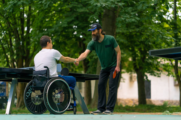Ένας ανάπηρος χωρίς αποκλεισμούς με μια ρακέτα στο χέρι κάνει χειραψία με έναν μεγαλύτερο πριν από ένα παιχνίδι πινγκ πονγκ ενάντια στο τραπέζι του τένις στο παρασκήνιο - Φωτογραφία, εικόνα