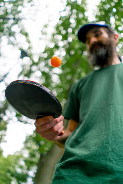 Un anciano junto a una mesa de ping pong azul golpea una pelota naranja en una raqueta de tenis en un parque de la ciudad - Foto, Imagen