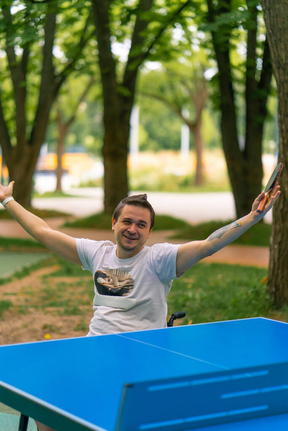 Egy inkluzív fogyatékos férfi ütővel a kezében ünnepli, hogy megnyerte a ping-pong játékot a városi park hátterében. - Fotó, kép