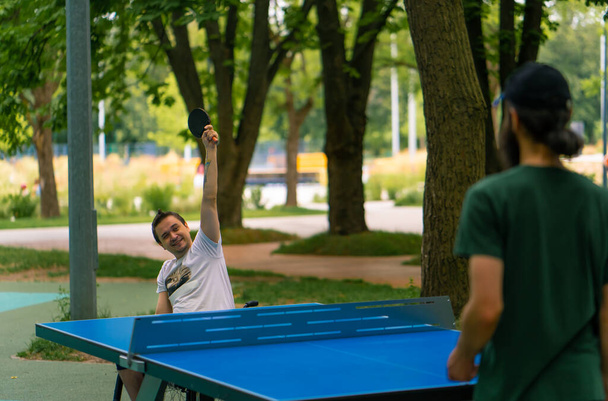 Inklusion glücklich Ein behinderter Mann im Rollstuhl spielt im Stadtpark Tischtennis gegen einen alten Mann mit grauem Bart - Foto, Bild