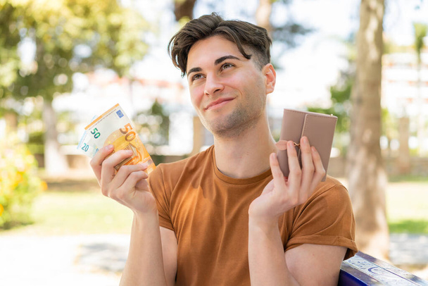 młody przystojny mężczyzna trzymając portfel z pieniądze i patrząc to - Zdjęcie, obraz