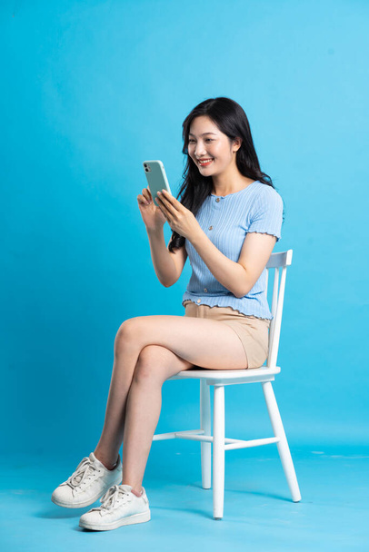 Portret pięknej Azjatki siedzącej na krześle, na niebieskim tle - Zdjęcie, obraz