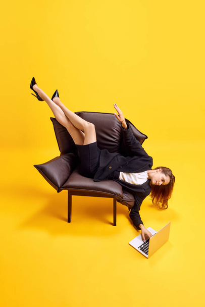 Young business woman lying on armchair in uncomfortable position, working on laptop against bright yellow background. Concept of business, working routine, deadlines, freelance, office, ad - 写真・画像