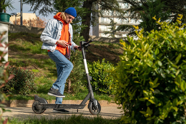 Junger Hipster steigt nach Aktivierung auf gemieteten Roller. Öko-Transport in der Stadt, Bezahlung für Roller durch Anwendung im Smartphone. Konzept moderner urbaner umweltfreundlicher Verkehr - Foto, Bild