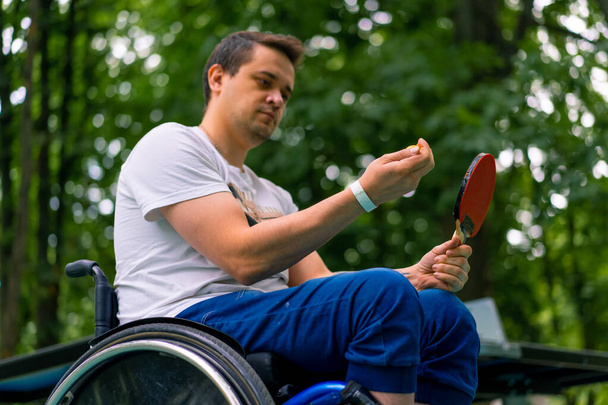 Inclusividade Um homem deficiente em uma cadeira de rodas com uma raquete e uma bola de pingue-pongue em suas mãos perto de uma mesa de tênis no parque - Foto, Imagem