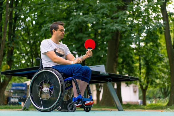 Ένταξη Ένας ανάπηρος σε αναπηρική καρέκλα παίζει πινγκ πονγκ σε ένα πάρκο της πόλης με φόντο τα δέντρα - Φωτογραφία, εικόνα