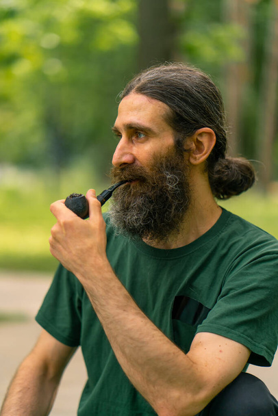 Porträt eines alten Mannes mit langen Haaren und grauem Bart, der im Stadtpark vor dem Hintergrund von Bäumen eine Pfeife raucht - Foto, Bild