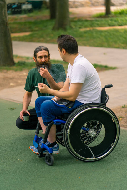 Ένταξη Ένας άνδρας με αναπηρία αλληλεπιδρά με έναν ηλικιωμένο άνδρα που καπνίζει πίπα σε ένα πάρκο της πόλης με φόντο τα δέντρα - Φωτογραφία, εικόνα