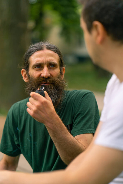 Inclusividad Un hombre con discapacidad interactúa con un anciano fumando una pipa en un parque de la ciudad contra el telón de fondo de los árboles - Foto, imagen