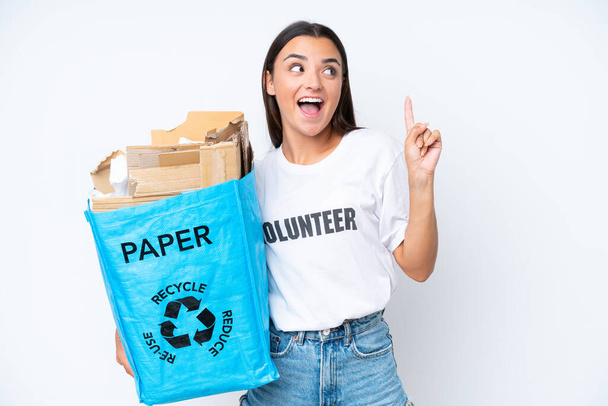 Giovane donna caucasica in possesso di un sacchetto di riciclaggio pieno di carta per riciclare isolato su sfondo bianco con l'intenzione di realizzare la soluzione mentre solleva un dito - Foto, immagini