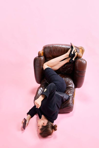 Female employee sitting on fallen armchair and looking on mobile phone against pink studio background. Chatting. Concept of business, working routine, deadlines, freelance, office, ad - 写真・画像