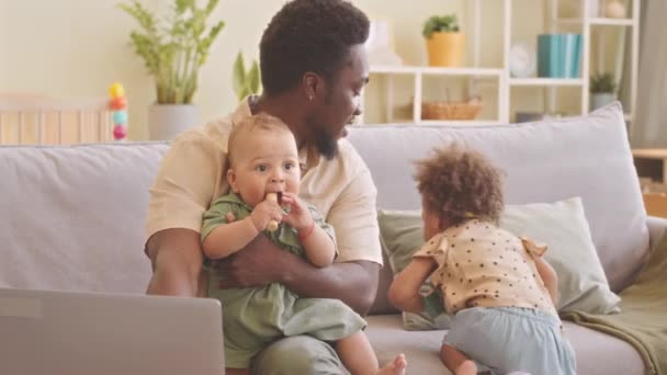 Young African American father of two toddlers trying to work on laptop from home while taking care of his kids - Filmmaterial, Video