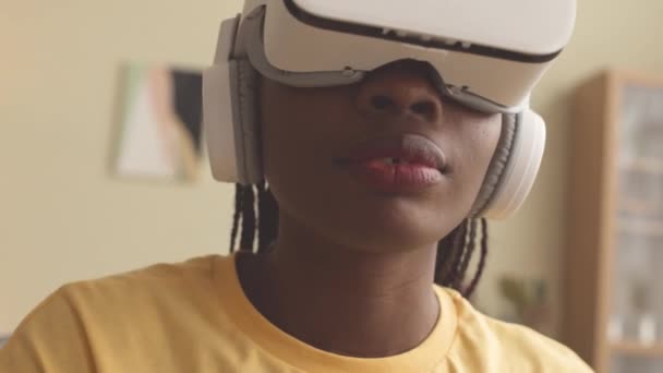 Medium close-up van geconcentreerde jonge zwarte vrouw in vr headset met behulp van vr gaming controller tijdens het spelen van vr spel op de bank in de woonkamer - Video
