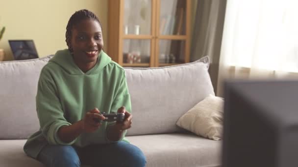 Fotografía media de una joven afroamericana en sudadera con capucha de gran tamaño usando un controlador de juego mientras juega videojuegos en casa - Imágenes, Vídeo