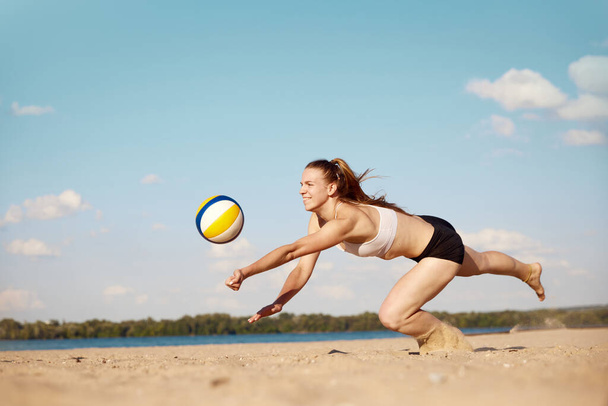 Competitive young woman athlete playing beach volleyball, hitting ball with hands and falling down on sand. Match. Concept of sport, active and healthy lifestyle, hobby, summertime, ad - Φωτογραφία, εικόνα