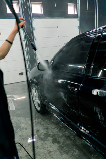 Ένας αρσενικός υπάλληλος πλυντηρίου αυτοκινήτων πλένει ένα μαύρο πολυτελές αυτοκίνητο με πλυντήριο υψηλής πίεσης στο πλυσταριό αυτοκινήτων - Φωτογραφία, εικόνα